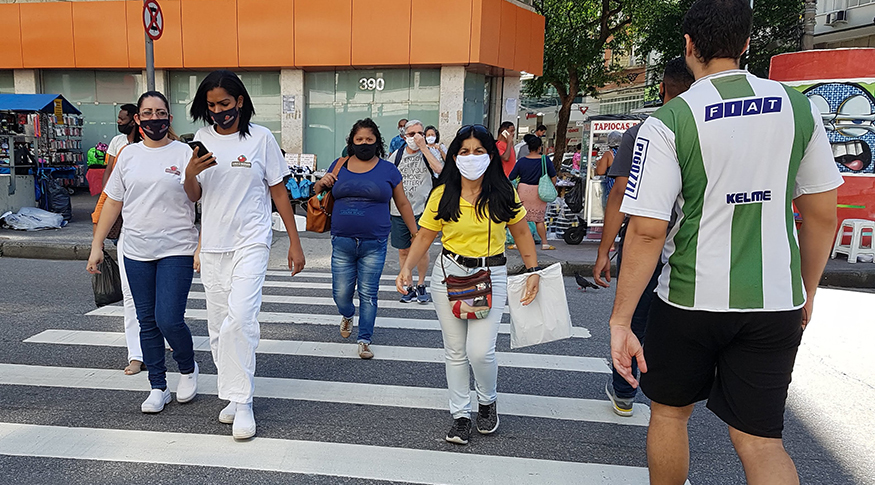 #PraCegoVer Pedestres usando máscaras de proteção, atravessando uma grande rua da cidade.