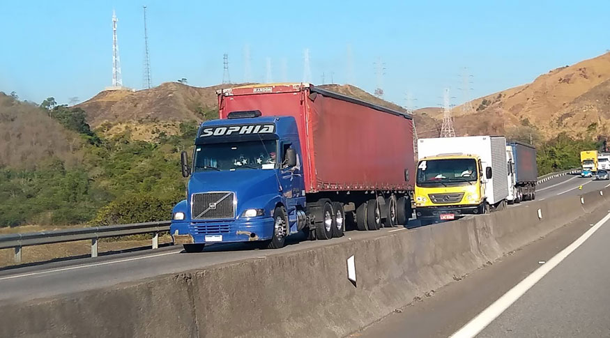 #pracegover A foto mostra um trecho de uma rodovia. Em primeiro plano aparece um caminhão com a frente azul e o resto em vermelho. Logo atrás, aparece outro caminhão, com a frente amarela e o restante prateado. Ao fundo, se vê mais alguns caminhões.