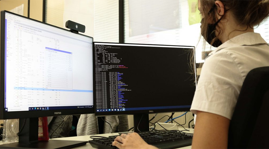 #pracego ver Uma pessoa do lado direito da imagem, sentada numa mesa de trabalho, usando duas telas de computador.