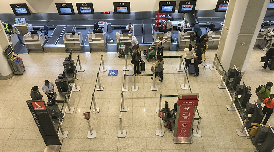 #PraCegoVer A foto mostra um salão de aeroporto visto de cima, com pessoas carregando malas e guichês de companhias aéreas.