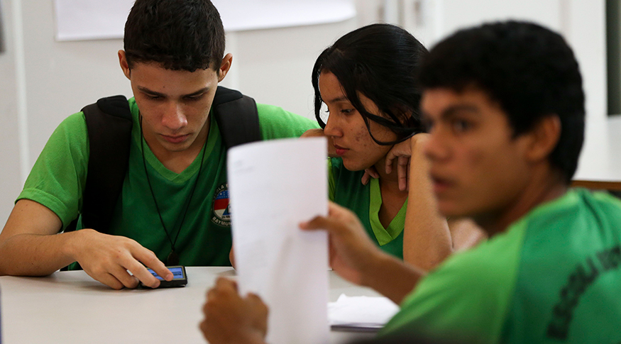#PraCegoVer A foto mostra três jovens uniformizados, estudando juntos