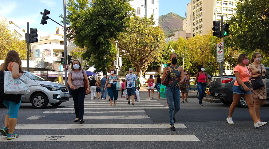 #PraCegoVer A foto mostra pessoas atravessando uma rua, na faixa de pedestres e ao fundo, muitas árvores.