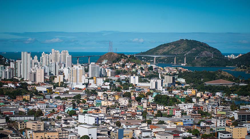 A imagem mostra uma vista panorâmica da cidade de Vitória/ES, destacando o contraste entre a paisagem litorânea e a área urbana da capital capixaba.