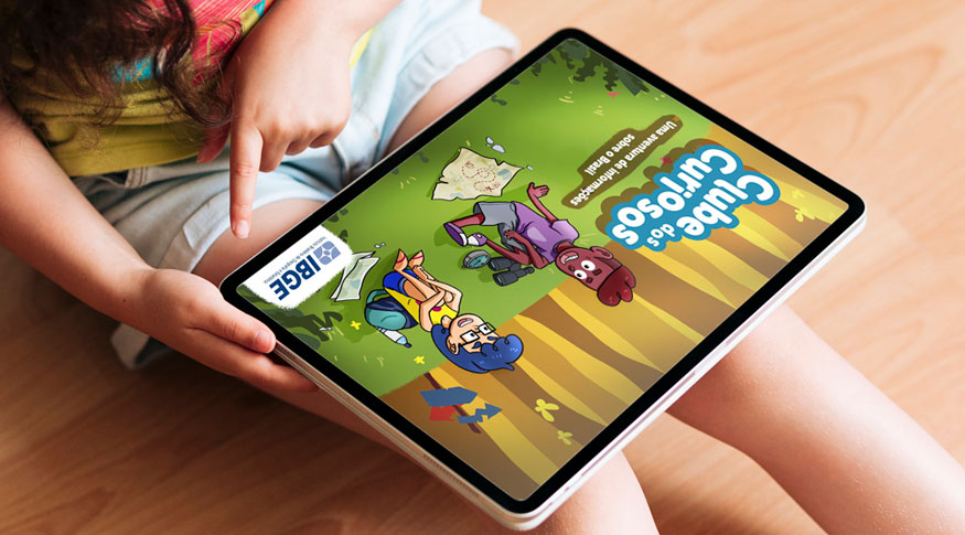 #PraCegoVer Vista de cima uma criança sentada com um tablet sobre as pernas. O tablet exibe a capa do livro e uma das mãos aponta para o canto direito onde está localizada o logotipo do IBGE na capa do livro 