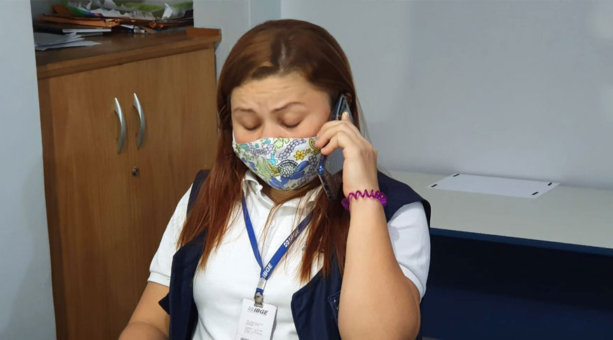 #PraCegoVer Mulher usando máscara estampada, blusa branca, colete azul escuro e crachá falando no celular