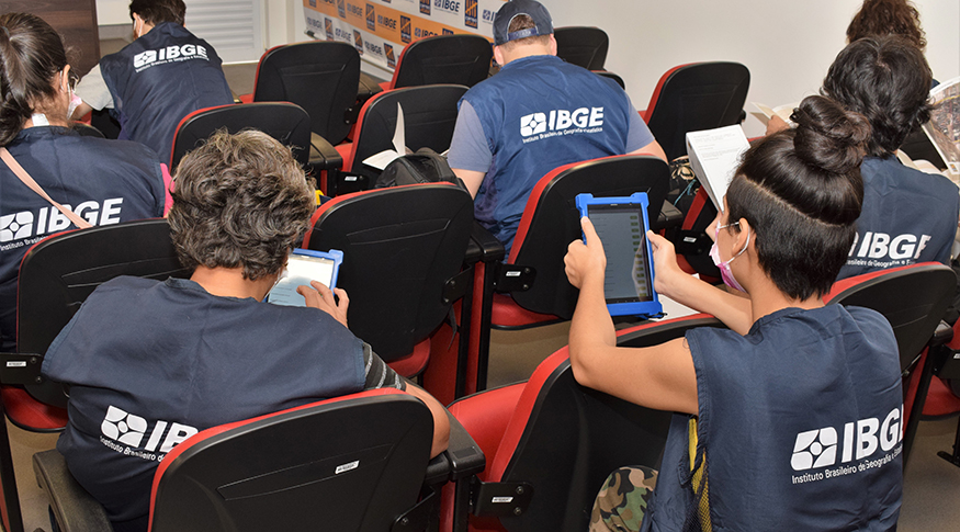 #PraCegoVer A foto mostra algumas pessoas, vestidas com o colete azul do IBGE, em uma sala de aula com cadeiras vermelhos, todas segurando um Dispositivo Móvel de Coleta.