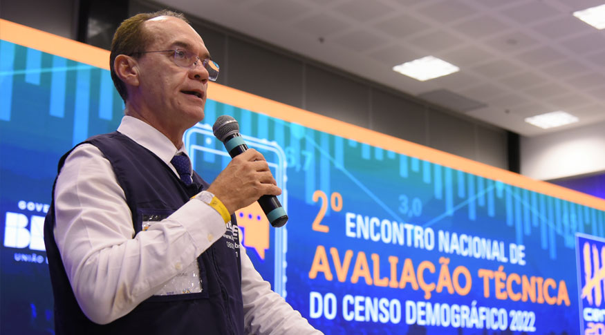 #Paratodoslerem A imagem tem um homem localizado do lado esquerdo segurando um microfone. Ele veste camisa social branca de manga comprida e colete azul escuro, uniforme do IBGE. Do centro para a dieita da foto o baner do evento projetado no telão